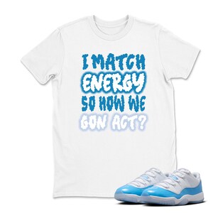 May include: White t-shirt with the text "I MATCH ENERGY SO HOW WE GON ACT?" in blue cloud-like font. A pair of white and light blue sneakers are in the foreground. The t-shirt and sneakers are likely part of a streetwear or casual fashion ensemble.