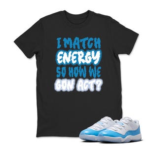May include: Black t-shirt with the text "I MATCH ENERGY SO HOW WE GON ACT?" in blue and white. A pair of light blue and white sneakers are in front of the shirt.