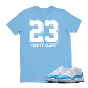 Puede incluir: Una camiseta azul claro con el n&uacute;mero 23 en blanco y la frase "KEEP IT CLASSIC". Un par de zapatillas deportivas azules y blancas est&aacute;n delante de la camiseta.