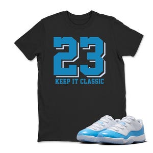 Puede incluir: Camiseta negra con el n&uacute;mero 23 en azul y blanco, con el texto "KEEP IT CLASSIC". Un par de zapatillas deportivas azules y blancas est&aacute;n delante de la camiseta.