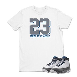 May include: White t-shirt with the number 23 in gray and blue, with the text "KEEP IT CLASSIC" below. A pair of gray, white, and black sneakers with blue laces are in the foreground. The sneakers are a high-top style.
