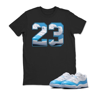 May include: Black t-shirt featuring the number "23" in a light blue and white design, with a pair of matching light blue and white sneakers in the foreground. The shirt's graphic incorporates an image of the sneakers.