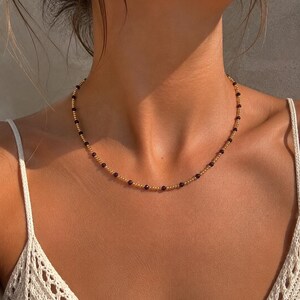 May include: A delicate beaded necklace featuring a repeating pattern of small, metallic gold-toned spherical beads interspersed with single, dark red, faceted gemstone beads, creating a refined and symmetrical circular design around the neck.