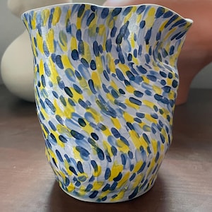 May include: A ceramic vase with a wavy, irregular rim and a white base color. The surface is covered in a dense pattern of small, hand-painted brushstrokes in shades of bright yellow, light blue, and dark navy blue, creating a textured appearance.