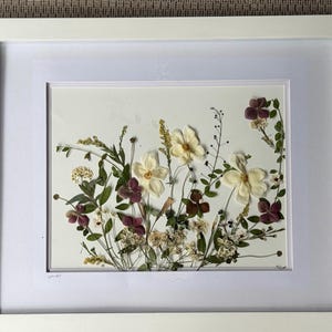 May include: A framed botanical art piece featuring pressed flowers. The arrangement includes cream-colored flowers, deep burgundy blooms, and various green foliage. The artwork is set against a white background and framed in a white wooden frame.