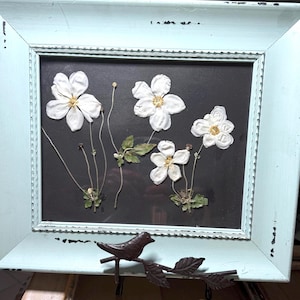 Puede incluir: Arreglo floral enmarcado con flores blancas con centros amarillos sobre un fondo negro. El marco es azul claro desgastado, y un pájaro decorativo de metal está posado debajo.