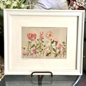 May include: A framed artwork featuring pressed flowers in shades of pink, white, and yellow. The flowers are arranged against a neutral background and encased in a white frame. The artwork is displayed on a glass table.