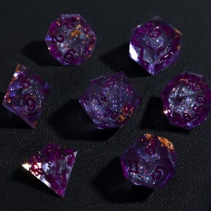 Könnte beinhalten: Sieben violette, polyedrische Würfel mit Glitzer und Goldflocken. Die Würfel sind transparent mit dunkelvioletten Zahlen. Die Würfel sind auf einer schwarzen Oberfläche angeordnet.
