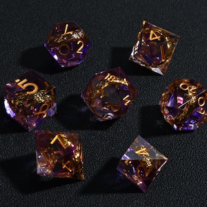May include: A collection of seven polyhedral dice with a purple and gold color scheme. The dice are transparent, with gold-colored numbers and internal designs. The dice are arranged on a dark surface, suitable for tabletop gaming.
