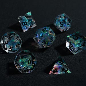 Könnte beinhalten: Sieben polyedrische Würfel für Tabletop-Spiele. Die transparenten Würfel haben schillernde blaue und grüne Einschlüsse und schwarze Zahlen. Enthält einen W4, W6, W8, W10, W12, W20 und einen Prozentwürfel.