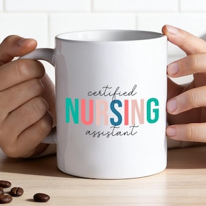Könnte beinhalten: Weiße Keramik-Tasse mit den Worten "certified NURSING assistant" in Türkis, Rosa und Blau. Die Tasse wird von einer Person gehalten. Kaffeebohnen und ein Zweig Grünzeug liegen auf der Holzoberfläche.