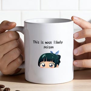 Könnte beinhalten: Weiße Keramik-Tasse mit dem Text "This is most likely poison" und einer Cartoon-Illustration einer Figur mit tealfarbenem Haar und einer blauen Schleife. Die Tasse wird von einer Person gehalten. Kaffeebohnen und ein Zweig Grünzeug liegen auf dem Tisch.
