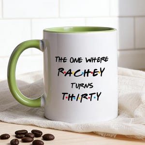 Könnte beinhalten: Weißer Keramikbecher mit grünem Henkel und Innenraum, mit dem Text "The One Where Rachey Turns Thirty" in einer bunten Schriftart. Der Becher steht auf einem weißen Tuch auf einer Holzoberfläche.