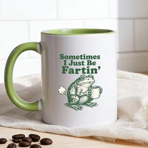 Könnte beinhalten: Weißer Keramikbecher mit grünem Henkel und Innenraum, mit dem Text "Sometimes I Just Be Fartin'" und einem Cartoon-Frosch, der eine Tasse hält. Der Becher steht auf einem weißen Tuch, mit Kaffeebohnen und einem Zweig Grünzeug in der Nähe.