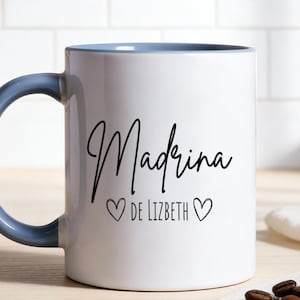 Könnte beinhalten: Weißer Keramikbecher mit blauem Henkel und Rand. Der Becher trägt das Wort "Madrina" in schwarzer Schrift, darunter "de Lizbeth" und zwei Herzsymbole. Der Becher steht auf einer Holzoberfläche.