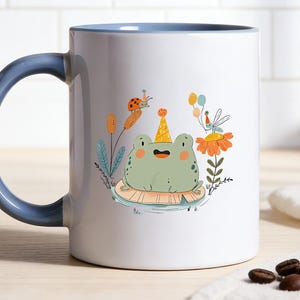 Könnte beinhalten: Weißer Keramikbecher mit blauem Griff und Innenrand. Der Becher zeigt eine skurrile Illustration einer grünen Frosches mit Partyhut, umgeben von Blumen und Insekten. Eine lustige, dekorative Kaffeetasse.