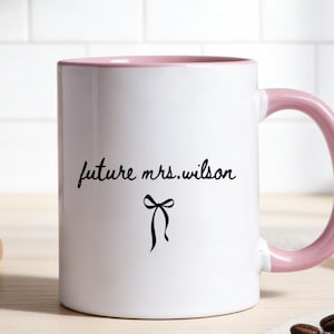 Könnte beinhalten: Weißer Keramikbecher mit rosa Innenraum und Henkel. Der Becher trägt den Text "future mrs. wilson" in handschriftlichem Stil, mit einer schwarzen Schleifenillustration darunter. Der Becher steht auf einer hellen Holzoberfläche.