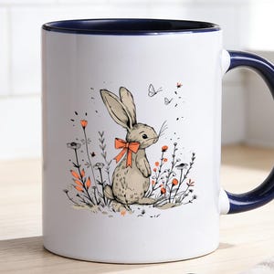 Könnte beinhalten: Weiße Keramiktasse mit marineblauem Innenraum und Griff. Die Tasse zeigt eine skurrile Illustration eines Hasen mit roter Schleife, umgeben von Blumen und Schmetterlingen. Kaffeebohnen und eine Leinen-Serviette liegen auf dem Tisch.