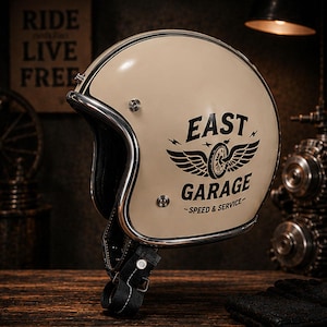 Può includere: Un casco da moto aperto color crema con bordo cromato. Il lato presenta una grafica nera di una ruota alata con il testo EAST GARAGE SPEED & SERVICE. Include un cinturino sottogola nero con fibbia in metallo e bottoni a pressione argentati.