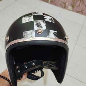 Könnte beinhalten: Ein mattschwarzer Jethelm für Motorräder mit einem abgenutzten schwarz-weißen Schachbrettmuster auf der Oberseite. Der Helm verfügt über eine metallisch silberne Zierleiste an der Vorderkante und drei Druckknöpfe zur Visierbefestigung.
