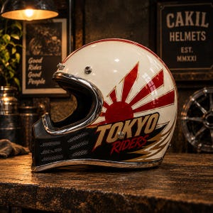 May include: A cream-coloured full-face motorcycle helmet with a red sunburst graphic and the text TOKYO RIDERS in gold and red lettering. The helmet features a black chin guard with silver mesh vents and a polished chrome trim along the visor opening.