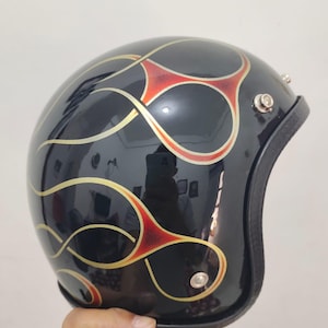 May include: A glossy black open-face motorcycle helmet featuring a decorative pinstripe design in gold and red. The helmet has a smooth, rounded shell with metallic silver snaps along the side for attaching accessories and a black trim edge.