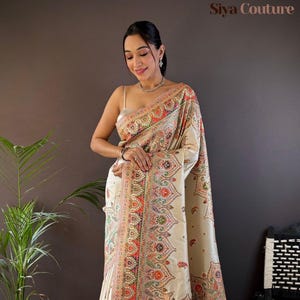 May include: Ivory-colored sari with intricate paisley and floral patterns in red, orange, green, and pink. The border features detailed designs and tassels. The sari is draped over a person, showcasing the elegant textile.