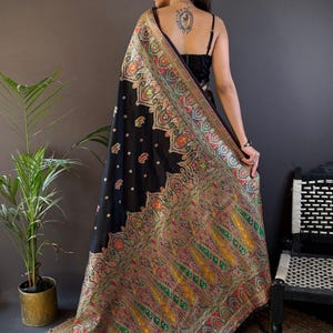 May include: A black sari with intricate, colorful embroidery. The sari features a detailed paisley pattern in shades of red, green, and gold. The image shows the sari draped over a person's shoulders, with the text "Siya Couture" visible.