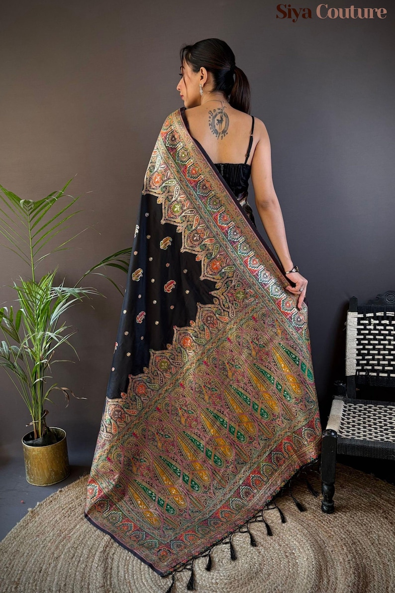 May include: A black sari with intricate, colorful embroidery. The sari features a detailed paisley pattern in shades of red, green, and gold. The image shows the sari draped over a person's shoulders, with the text "Siya Couture" visible.