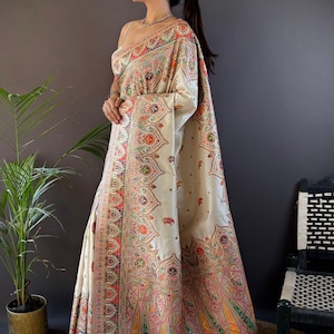 May include: Cream-colored sari with intricate paisley and floral patterns in shades of red, orange, green, and pink. The sari features a detailed border and the text "Siya Couture". The garment is draped over a person.