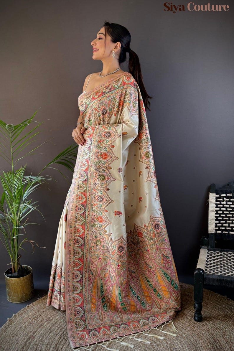 May include: Cream-colored sari with intricate paisley and floral patterns in shades of red, orange, green, and pink. The sari features a detailed border and the text "Siya Couture". The garment is draped over a person.
