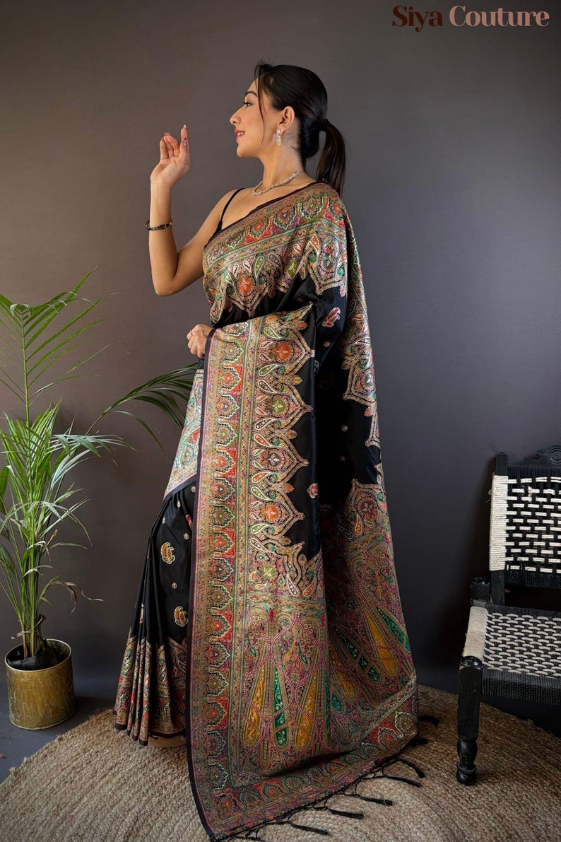 May include: A black sari with intricate, colorful embroidery. The sari features a detailed paisley pattern in shades of red, green, yellow, and orange. The garment is draped over a person, showcasing the design and texture of the fabric.