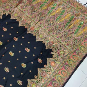 May include: Black shawl with intricate paisley and floral patterns in shades of yellow, green, pink, and orange. The border features a detailed design, and the edges are finished with black tassels. The shawl is a textile.