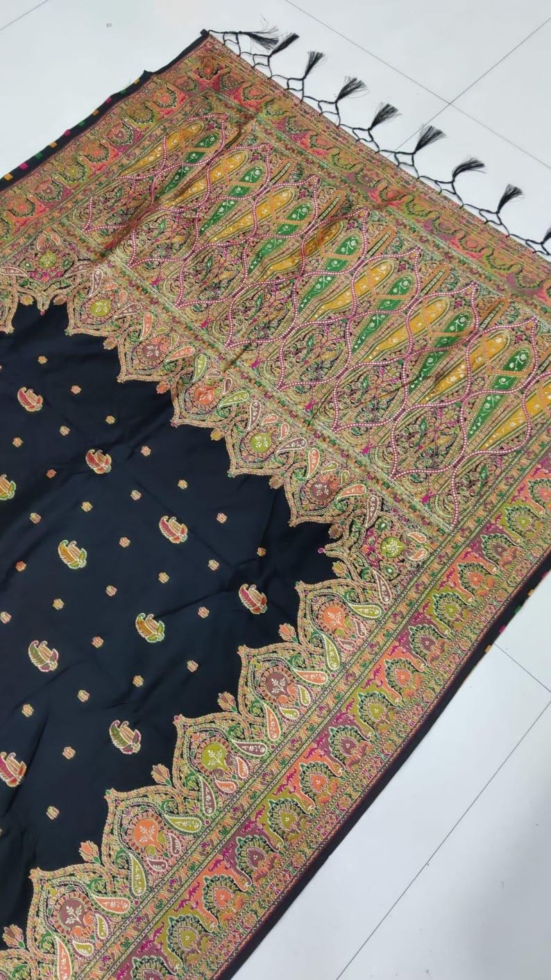 May include: Black shawl with intricate paisley and floral patterns in shades of yellow, green, pink, and orange. The border features a detailed design, and the edges are finished with black tassels. The shawl is a textile.