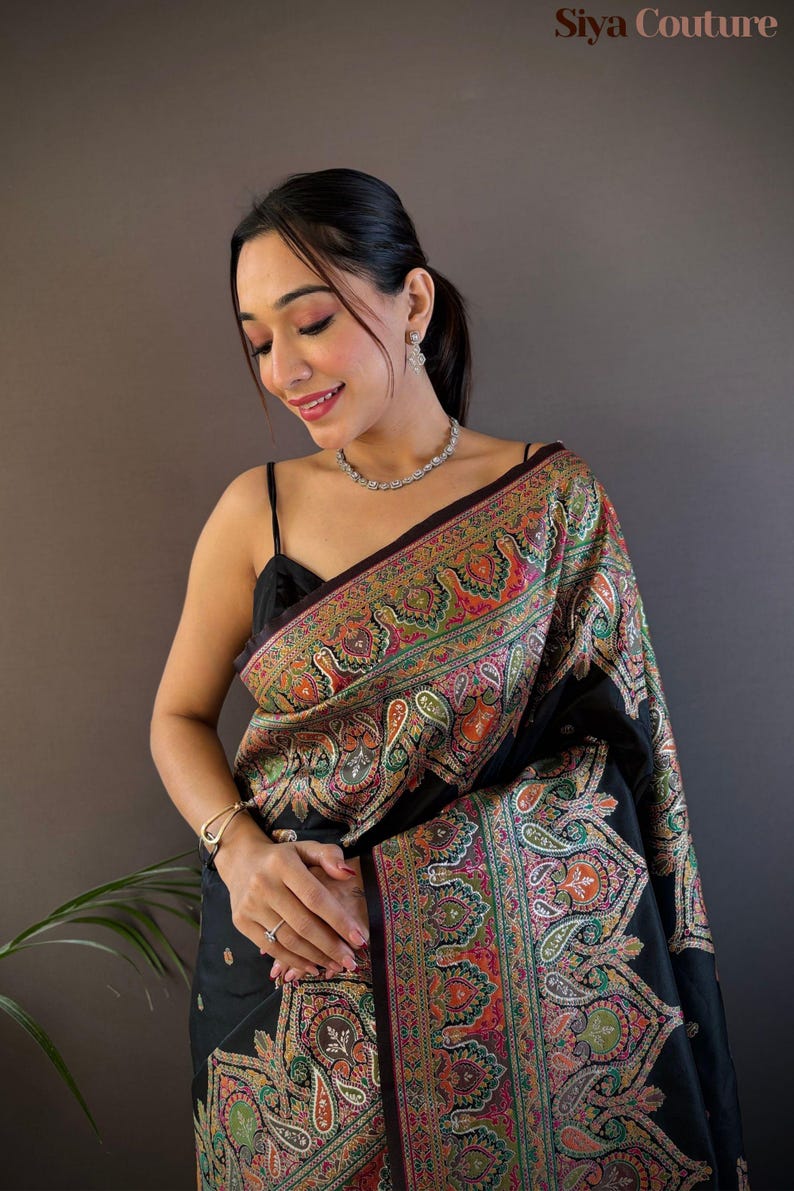 May include: Black sari with a colorful paisley pattern. The sari features a detailed design with shades of red, green, orange, and yellow. The sari is draped over the shoulders.