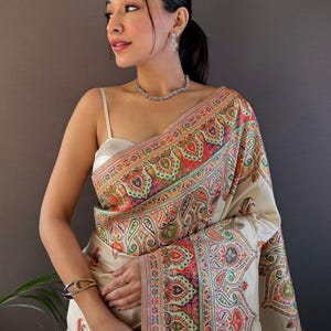 May include: A woman wearing a cream-colored sari with a vibrant paisley border. The sari features intricate patterns in shades of red, green, orange, and pink. The image also shows a silver necklace, earrings, and bracelet. The text "Siya Couture" is visible.