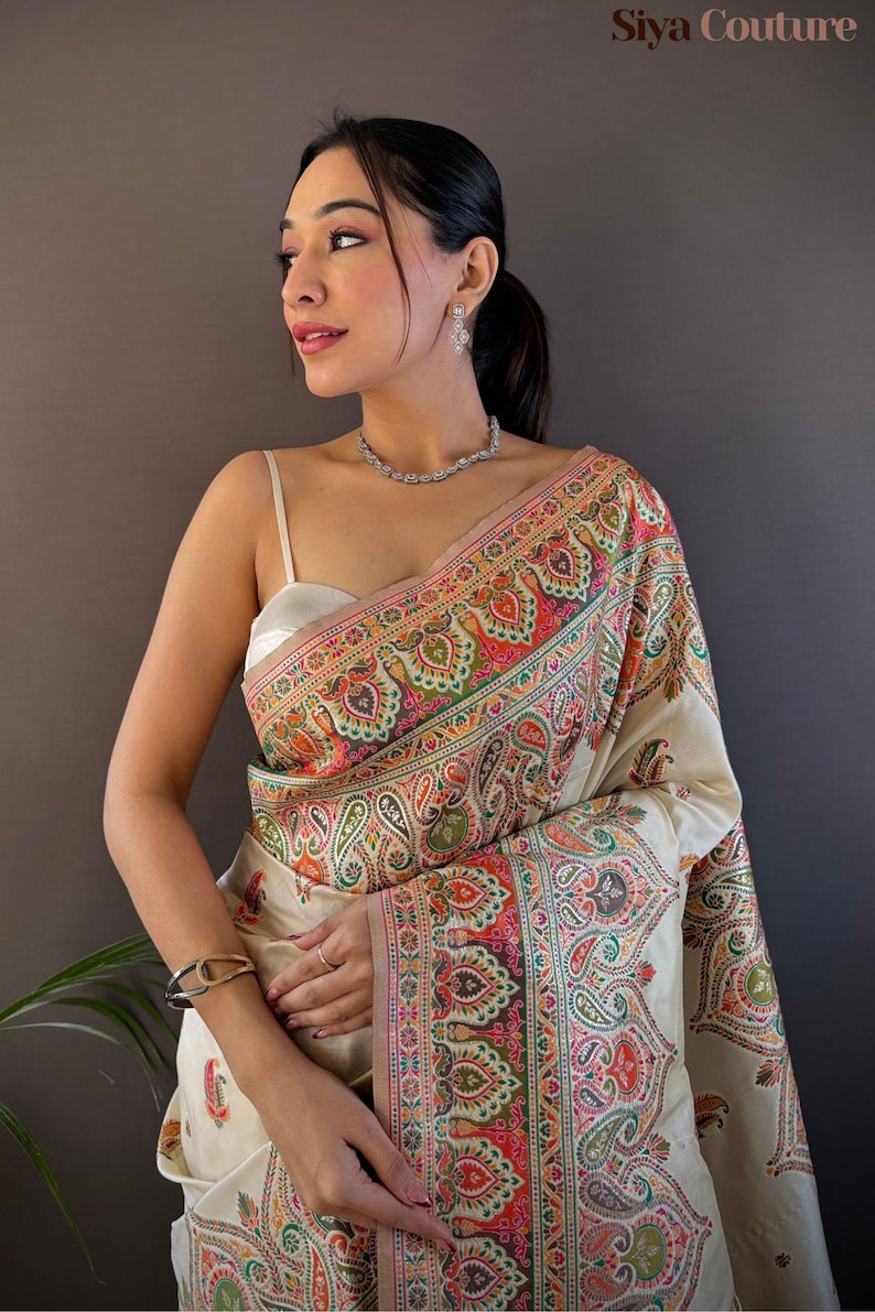 May include: A woman wearing a cream-colored sari with a vibrant paisley border. The sari features intricate patterns in shades of red, green, orange, and pink. The image also shows a silver necklace, earrings, and bracelet. The text "Siya Couture" is visible.