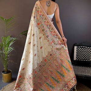 May include: Cream-colored shawl with intricate, colorful paisley patterns and fringe. The shawl features a detailed border with red, orange, green, and pink accents. The text "Siya Couture" is visible in the upper right corner. The shawl is draped over a person.