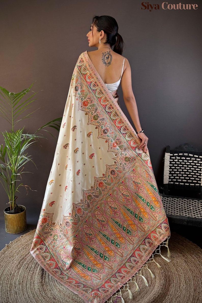 May include: Cream-colored shawl with intricate, colorful paisley patterns and fringe. The shawl features a detailed border with red, orange, green, and pink accents. The text "Siya Couture" is visible in the upper right corner. The shawl is draped over a person.