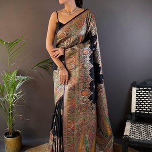 May include: A black sari with a colorful paisley pattern and a decorative border. The sari is draped over a person, with the text "Siya Couture" in the upper right corner. The sari has a fringe at the bottom.