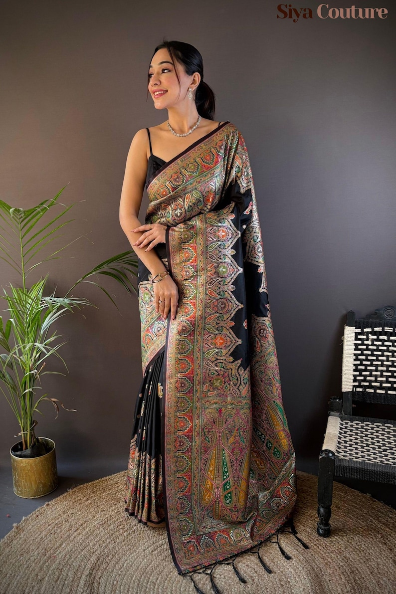 May include: A black sari with a colorful paisley pattern and a decorative border. The sari is draped over a person, with the text "Siya Couture" in the upper right corner. The sari has a fringe at the bottom.