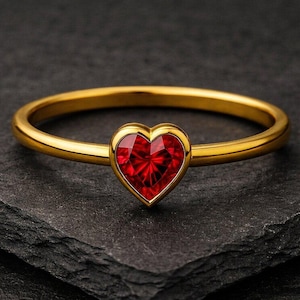 Puede incluir: Un anillo dorado con una gema roja facetada en forma de corazón. El anillo se encuentra sobre una superficie oscura y texturizada, resaltando la brillante banda dorada y el rojo vibrante de la gema. Un diseño simple y elegante.