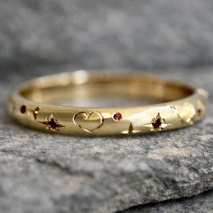 May include: A gold ring with celestial and heart-shaped cutouts and small red gemstones. The ring features a polished finish and is set against a gray stone surface. The design includes stars, hearts, and dots.
