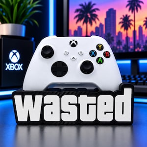 Könnte beinhalten: Ein weißer kabelloser Gamecontroller in einem schwarzen Ständer mit dem Wort WASTED in fetten weißen Großbuchstaben auf einem Hintergrund mit Carbonfaser-Textur. Der Controller verfügt über schwarze Analogsticks, ein Steuerkreuz und farbige Tasten.