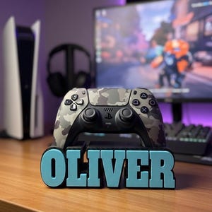 Könnte beinhalten: Ein Tarnmuster-Videospiel-Controller liegt auf einem Ständer mit dem Namen "OLIVER" in blauen Buchstaben. Eine Spielkonsole und Kopfhörer sind im Hintergrund zu sehen. Der Controller hat schwarze Tasten und eine schwarze Basis.