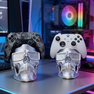 Könnte beinhalten: Zwei silberne Controller-Halter in Schädelform, jeweils mit einem Videospiel-Controller obenauf. Ein Controller hat ein Tarnmuster, der andere ist weiß. Die Halter haben ein metallisches Finish und stehen auf einem Schreibtisch.