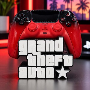 Könnte beinhalten: Ein rot-schwarzer Videospiel-Controller mit Farbverlauf auf einem schwarzen Ständer mit dem weißen Schriftzug GRAND THEFT AUTO und einem Sternsymbol. Der Controller hat rote Analogsticks, ein schwarzes Touchpad und schwarze Tasten mit roten Symbolen.
