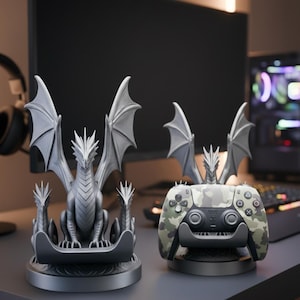 Könnte beinhalten: Zwei graue Controller-Ständer im Drachen-Design. Der linke Ständer zeigt einen zentralen Drachen mit ausgebreiteten Flügeln und zwei kleineren Drachenköpfen an den Seiten. Der rechte Ständer hält einen Camouflage-Controller mit einer Drachenfigur.