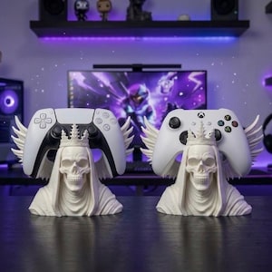 Könnte beinhalten: Zwei elfenbeinfarbene Controller-Ständer in Schädelform. Jeder Schädel trägt eine Krone und hat flügelartige Strukturen. Ein Ständer hält einen weißen PlayStation 5-Controller, der andere einen Xbox-Controller. Im Hintergrund ein Computer und Regale.