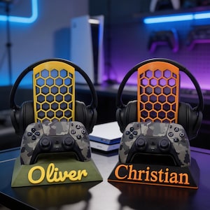 Könnte beinhalten: Zwei Gaming-Controller-Ständer mit passenden Kopfhörern. Ein Ständer ist grün mit dem Namen "Oliver" in Gelb, der andere ist schwarz mit "Christian" in Orange. Jeder Ständer hält einen Camouflage-Controller und ein schwarzes Headset.